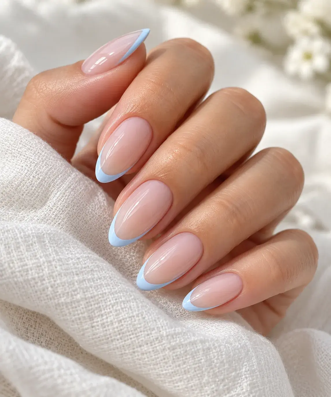 A hand showing almond-shaped nails with a glossy nude base and pale blue French tips resting on white fabric.