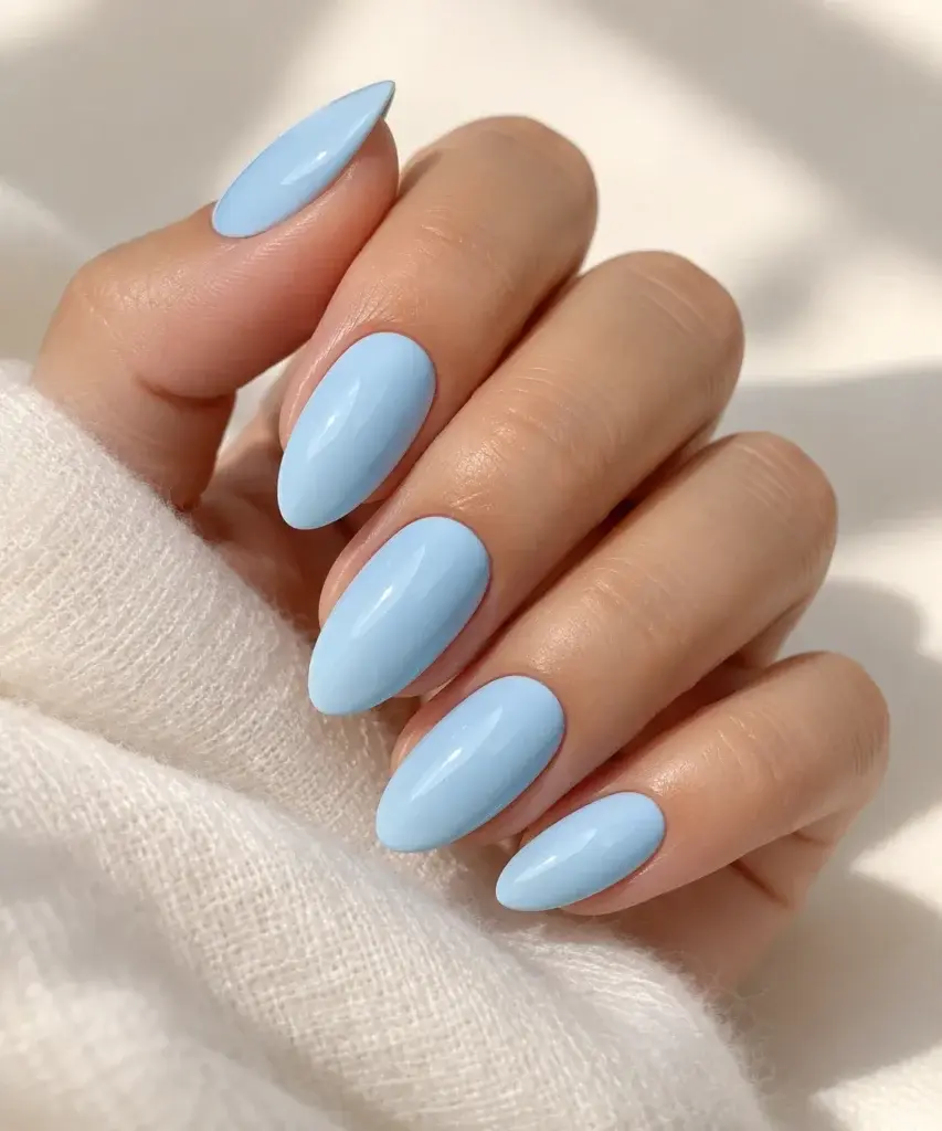 Hand with glossy oval-shaped nails painted in a soft sky blue polish resting against a white knit sweater.