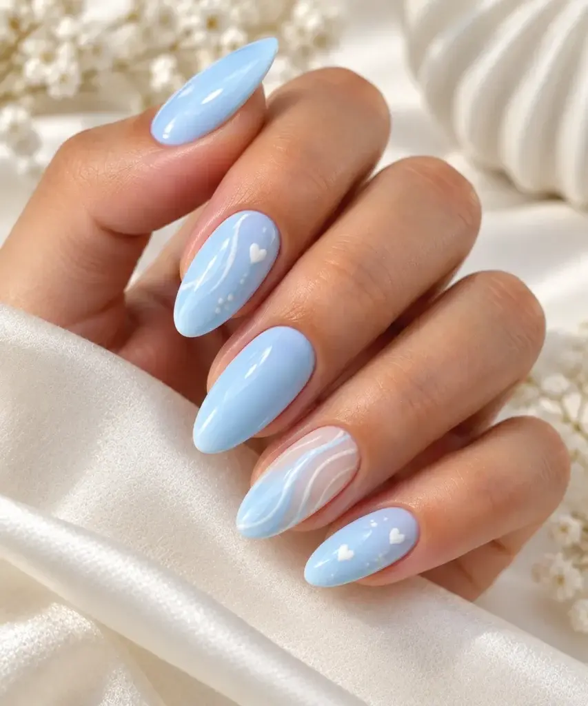 Close-up of a hand with long almond-shaped nails painted in soft light blue, featuring white heart and swirl designs, resting on white fabric.