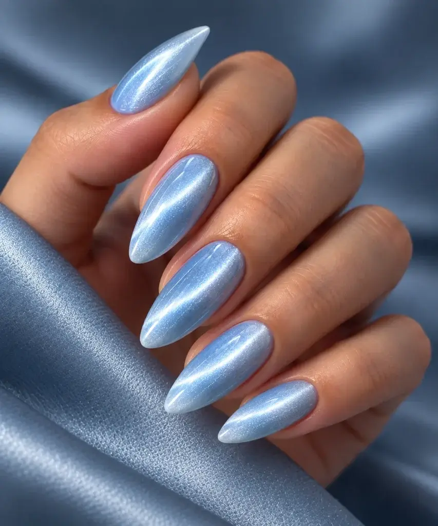 Close-up of long almond-shaped nails with a shimmering light blue cat eye finish, resting on a textured blue fabric.