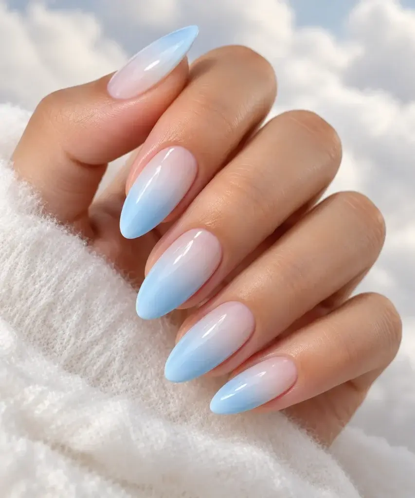 A hand with long, almond-shaped nails featuring a soft blue and white gradient polish, resting on a white fluffy surface against a cloudy sky background.
