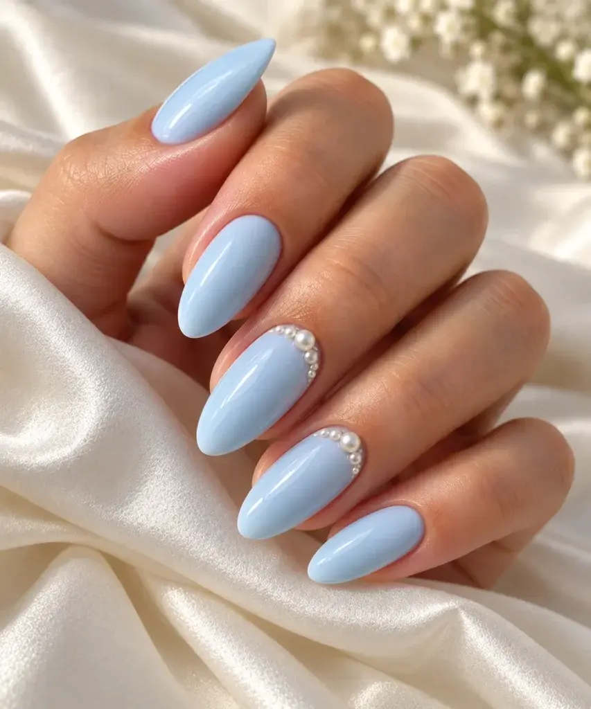 A hand with long, almond-shaped nails painted in a soft light blue shade, featuring small pearl details on the ring and middle fingers, resting on white satin fabric.